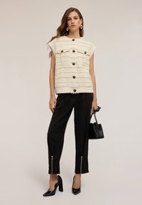 Donna che indossa un gilet senza maniche color crema con bottoni, pantaloni neri tapered con cerniere alla caviglia, tacchi neri e tiene una borsa nera.