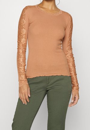 Kvinde iført en tætsiddende ribstrikket langærmet top med blomstrede blonder på ærmerne i terracottafarve, kombineret med olivengrønne bukser, stående foran en hvid baggrund.