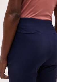 Κοντινό πλάνο ενός ατόμου που φοράει παντελόνι ψηλόμεσο σε ναυτικό μπλε χρώμα και πουκάμισο σε σκούρο ροζ απόχρωση, δείχνοντας την πίσω τσέπη και το κάτω μέρος του κορμού.