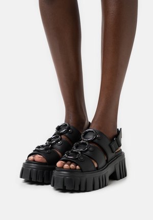 Sandales à plateforme noires avec semelles épaisses, sangles en cuir et accents métalliques ronds. Caractéristiques : bout ouvert et bride de cheville ajustable.