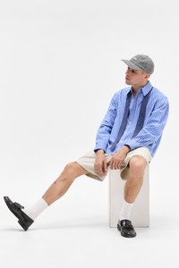 Ung mand iført en blå stribet skjorte, beige shorts, hvide sokker, sorte loafers og en ternet cap, siddende på en hvid kube mod en hvid baggrund.