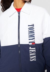 Tommy Jeans jacket features a navy and white color block design, zip-up front, classic collar, and embroidered logo on the side.