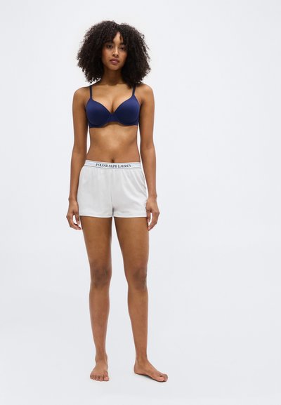 Γυναίκα με σγουρά μαλλιά που φοράει μπλε σουτιέν και λευκά σορτς Polo Ralph Lauren, όρθια ξυπόλητη μπροστά σε απλό φόντο.