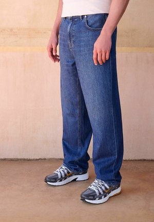 Person trägt lockere blaue Jeans und graue Sneaker mit weißen Schnürsenkeln und steht vor einem beigen Wand- und Bodenhintergrund.