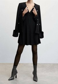Vestido negro con un delantero anudado, combinado con una chaqueta negra texturizada que tiene puños de pelo y botones decorados. Botines brillantes completan el look.