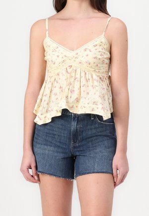 Mujer con blusa sin mangas amarilla con estampado floral y detalles de encaje, y shorts de mezclilla azul con dobladillo sin rematar, posando frente a un fondo liso.