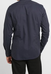 Mörkblå skjorta med knappar och lång ärm, en mjuk textur och en rundad fåll. Har detaljer med knappar vid ärmsluten.