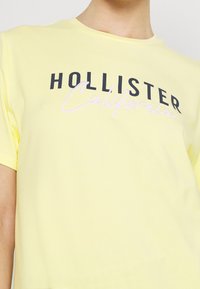 Żółta bawełniana koszulka z okrągłym dekoltem i krótkimi rękawami. Na przodzie znajduje się nadrukowany czarny i różowy napis: "HOLLISTER California."