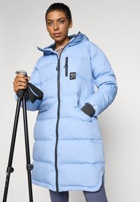 Giacca gonfiabile di colore azzurro chiaro con cappuccio, cerniera frontale, tasche laterali e dettagli neri. Il modello tiene in mano dei bastoni da trekking, evidenziando il design funzionale.
