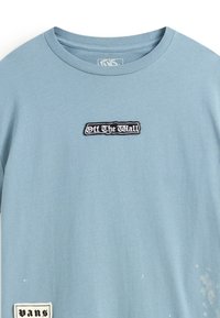 Camiseta de algodón de color azul claro con un parche bordado "Off the Wall" en blanco y negro, y una pequeña etiqueta "Vans" cosida en el lado.