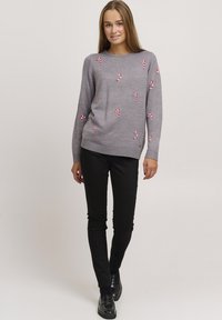 Siv sweater z rdečim in belim vezenjem v obliki paličic sladkarij, okrogla izrezana vrhnja, rebrasti manšeti in spodnji rob, kombiniran s črnimi hlačami in črnimi čevlji.