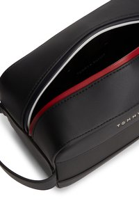 Μαύρο δερμάτινο νεσεσέρ Tommy Hilfiger με ανοιχτό λευκό φερμουάρ, κόκκινη εσωτερική λεπτομέρεια και λουράκι χειρός σε λευκό φόντο.