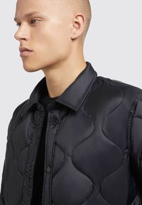 Sort quiltet jakke med et bølgemønster, trykknapper og krave. Stoffet fremstår glat og mat i tekstur.
