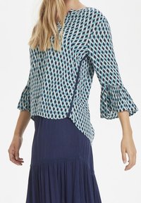 Top blu e bianco con maniche a campana, caratterizzato da un design geometrico. Il tessuto appare leggero con una vestibilità comoda.