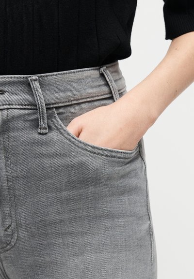 Χέρι που ακουμπάει μέσα στην μπροστινή τσέπη ψηλόμεσου γκρι τζιν, συνδυασμένο με μαύρο ριμπ πουκάμισο.