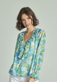 Blaugrüne Bluse mit Rüschenausschnitt, ausgestattet mit einem Aquarelldruck in Gelb und Aqua. Lange Ärmel mit elastischen Bündchen und Knopfdetail.