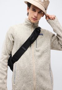 Νέος άνδρας που φοράει μπεζ φλις μπουφάν, μαύρη τσάντα χιαστί και μπεζ καπέλο τύπου bucket, ρυθμίζοντας το καπέλο με το ένα χέρι.