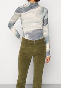 Persoon in een aansluitend, lang mouwtop met grijze en witte wolkachtige patronen en olijfgroene corduroy broek, staande met een hand op de heup.