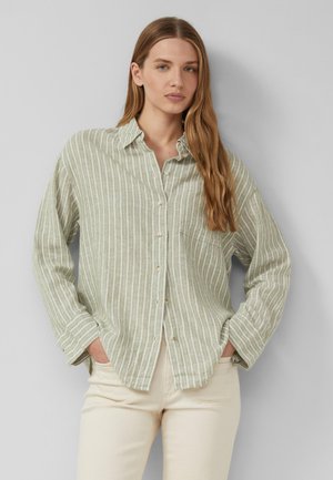 Vrouw met lang lichtbruin haar, gekleed in een lichtgroen-wit verticaal gestreept blouse met knopen en beige broek, staat met handen in de zakken.
