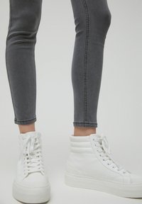 Zapatillas altas blancas con un acabado texturizado, suela plana y diseño de cordones, combinadas con unos jeans gris ajustados que tienen una costura lateral.
