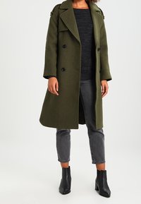 Dvojřadový olivově zelený vlněný kabát s velkými černými knoflíky, širokými klopami a ramenními epolety, nošený s tmavým svetr a šedými džíny.