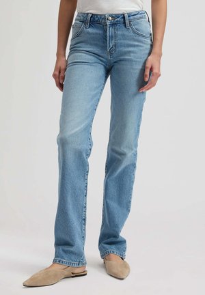 Model purtând blugi drepti de culoare albastru deschis, cu buzunare frontale și pantofi bej cu vârf ascuțit, stând pe un fundal simplu și deschis.