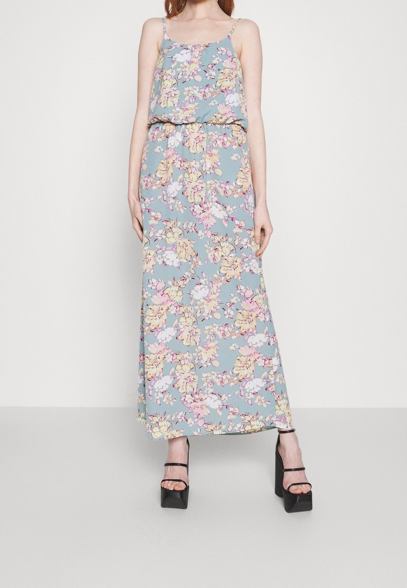 Femme portant une robe longue sans manches bleue à motifs floraux avec des bretelles spaghetti et des talons compensés noirs sur un fond uni.