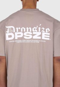 Beige Baumwoll-T-Shirt mit kurzen Ärmeln, das auf der Rückseite ein großes, weißes grafisches Textdesign mit "DPSZE" und Logodetails zeigt.