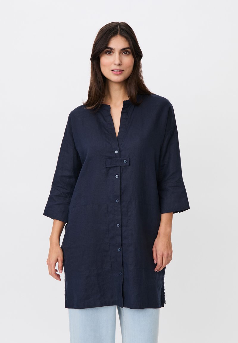 Femme portant une tunique à manches longues bleu marine boutonnée avec un col cranté et un pantalon bleu clair, debout devant un fond blanc uni.