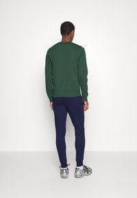 Sudadera verde con puños acanalados, combinada con joggers ajustados de color marino. Zapatillas deportivas grises con acentos amarillos y detalles en negro.