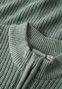 Nahaufnahme des gerippten Kragens und des texturierten Musters eines grünen Strickpullis, mit sichtbarem Stoffgewebe und Kleidungsetiketten im Ausschnitt.
