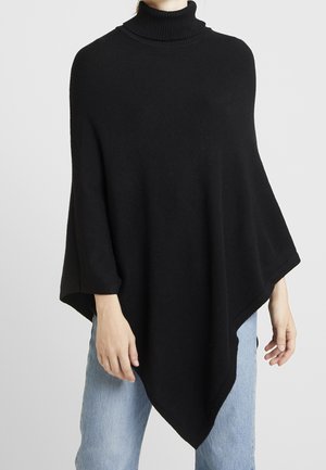 Femme portant un poncho en maille noire avec un col roulé, associé à un jean bleu clair, sur un fond gris uni.