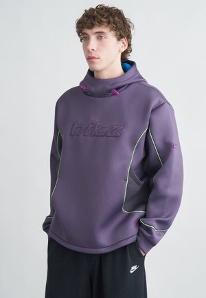 Sweat à capuche violet avec des accents verts néon contrastants, logo en relief, cordons de serrage et panneaux latéraux en maille. Associé à un pantalon noir arborant un logo.