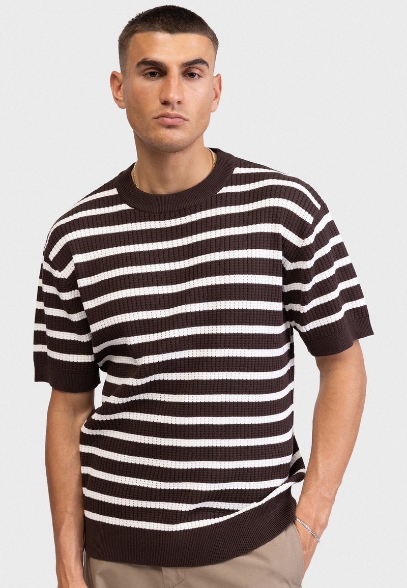 Νεαρός άνδρας με κοντά μαλλιά, φορεμένος με καφέ και λευκό ριγέ κοντομάνικο πουλόβερ και μπεζ παντελόνι, με το ένα χέρι στην τσέπη.