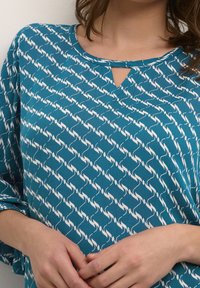 Kvinde iført en teal farvet langærmet top med et hvidt gentaget abstrakt mønster og en lille nøglehulsudskæring, hænderne blidt samlet om livet.