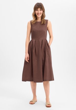 Vrouw glimlachend, staand, draagt een mouwloze bruine midi-jurk met plooien en beige sandalen, tegen een effen lichte achtergrond.