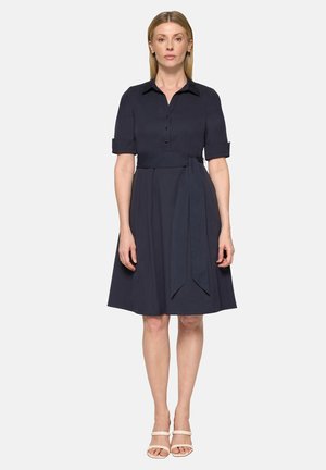 Frau steht mit dem Gesicht nach vorne und trägt ein knielanges, marineblaues Hemdblusenkleid mit kurzen Ärmeln und einem Taillengürtel, kombiniert mit weißen Sandalen mit offenem Zehenbereich.