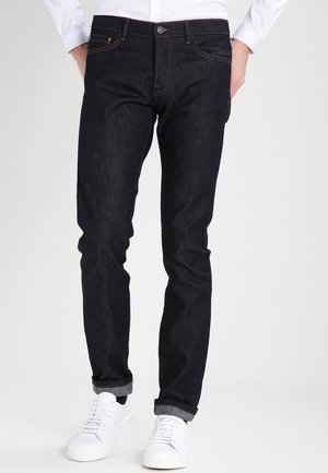 Mężczyzna w dopasowanych ciemnoniebieskich dżinsach ze zmarszczonymi mankietami i białych trampkach, z rękami w kieszeniach, stojący na jednolitym białym tle.