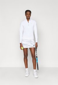 Balta ilgomis rankovėmis palaidinė su ketvirtadalio užtrauktuku ir logotipu, derinama su šviesiai pilku kamufliažo rašto sijonu. Laiko tenisą kamuoliuką ir raketę.