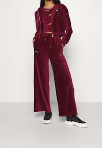 Mujer vistiendo pantalones anchos de terciopelo burdeos, sudadera corta con cremallera y top corto a juego, combinados con zapatillas negras y blancas.