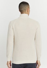 Beige neulepusero, jossa on korkea kaulus, resorit hihansuissa ja helmassa sekä teksturoitu kuvio. Takana näkyvät saumat ja istuva siluetti.