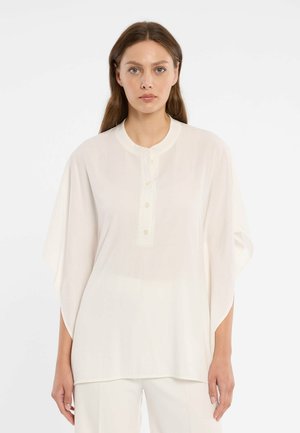 Vrouw met lang bruin haar, gekleed in een losse witte blouse met knopen en een witte broek, staand tegen een effen witte achtergrond.