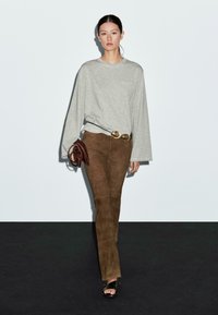 Vrouw die loopt in een losse grijze lange mouw shirt, bruine suède broek, zwarte sandalen, met een bruine clutch en een riem met grote gouden gespen.