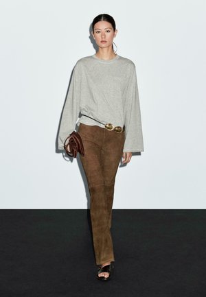 Woman walking wearing a loose gray long-sleeve shirt, brown suede pants, black sandals, holding a brown clutch bag and a belt with large gold buckles.