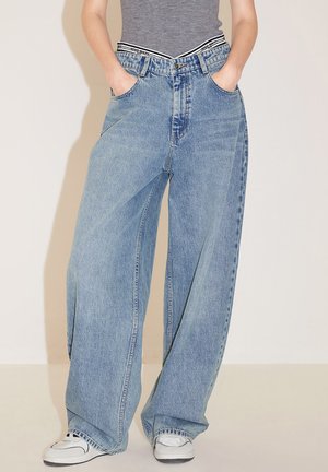 Jeans in denim blu a gamba larga con vita alta, lavaggio chiaro, doppi taschini laterali e vita a righe in contrasto. Sneakers bianche completano il look.
