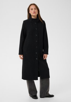 Donna in piedi con un lungo cappotto nero abbottonato con collo alto, indossa jeans grigi a gamba larga e scarpe nere, rivolta in avanti contro uno sfondo semplice.
