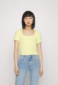 Top amarelo com nervuras e mangas curtas, com decote quadrado. O tecido parece suave e justo, combinado com jeans de cintura alta em azul claro.