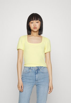 Top amarelo com nervuras e mangas curtas, com decote quadrado. O tecido parece suave e justo, combinado com jeans de cintura alta em azul claro.