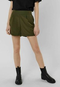 Krótkie spodenki w oliwkowej zieleni z wysokim stanem, plisowanym przodem i podwiniętym brzegiem, zestawione z czarnymi botkami za kostkę. Gładka faktura, casualowy krój.