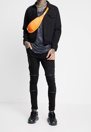 Giacca di jeans nera, t-shirt camouflage, jeans skinny neri strappati e sneakers nere con accenti riflettenti. Marsupio arancione come accessorio.